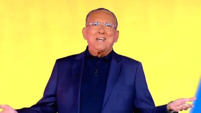Galvão Bueno sorri de óculos e braços abertos, vestindo paletó azul escuro sobre camisa polo preta, com fundo amarelo vibrante e desfocado