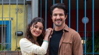 Gerluce (Sophie Charlotte) e Paulinho (Romulo Estrela) sorriem juntos na novela Três Graças, ela veste blusa clara e ele camisa marrom aberta no cenário