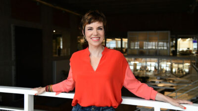 Gloria Vanique sorri apoiada em corrimão interno da Fundação Cásper Líbero usando blusa vermelha em ambiente corporativo moderno