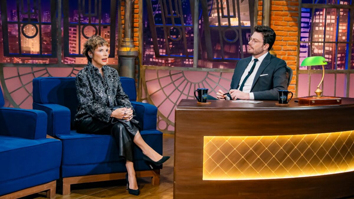 Glória Vanique veste jaqueta brilhante e calça escura sentada em poltrona azul conversando com Danilo Gentili de terno e óculos no cenário do The Noite