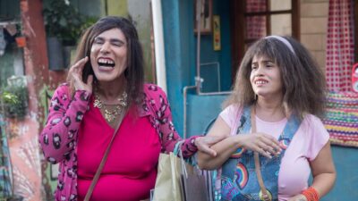 Rodrigo Sant'anna como Graça, rindo com sacolas de compras, e Isabelle Marques como Briti, de jardineira jeans, em cena de Tô de Graça - O Filme