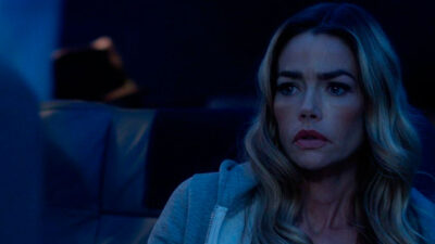 Denise Richards como Gretchen Blair no filme Altitude, a atriz olha fixamente para o lado com expressão séria, veste moletom cinza, fundo azulado