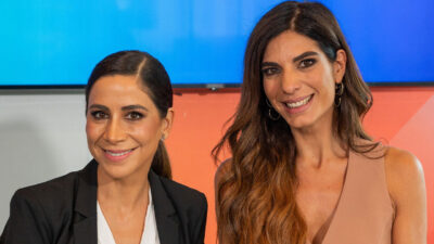 Jornalistas Julia Duailibi e Andréia Sadi sorriem juntas em estúdio com fundo azul e laranja Duailibi usa blazer preto e Sadi blusa bege sem manga