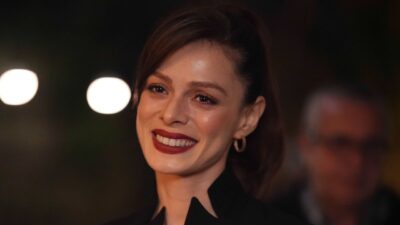 Atriz Ozge Ozpirincci como Karsu na novela Coração de Mãe sorri com batom vermelho intenso, cabelos presos, brincos de argola e traje preto elegante