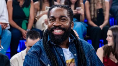 Lázaro Ramos sorri com dreadlocks longos e barba, vestindo jaqueta jeans azul sobre camiseta branca, em estúdio de televisão com plateia ao fundo