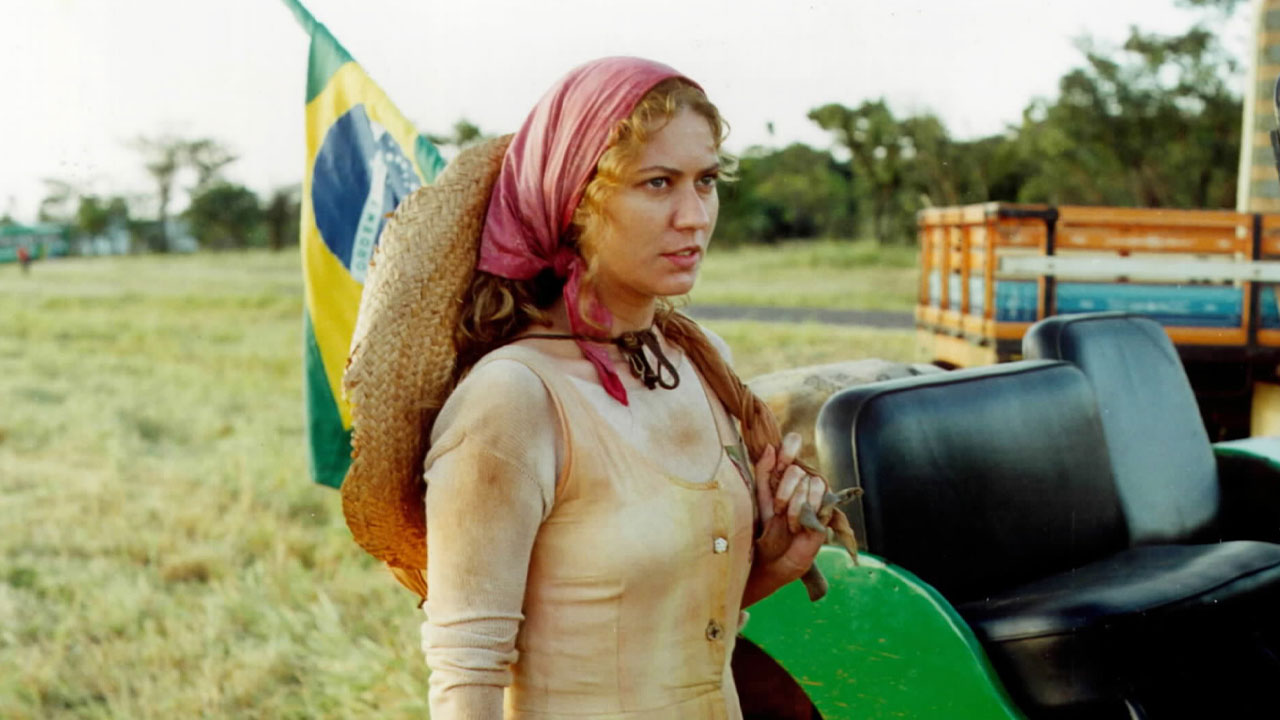Patrícia Pillar como Luana em O Rei do Gado com lenço vermelho na cabeça e chapéu de palha em cenário rural com bandeira do Brasil ao fundo