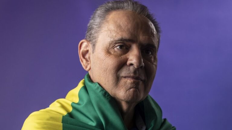 Luis Roberto olha para o lado com expressão serena, envolto em uma bandeira do Brasil sobre os ombros, contra um fundo roxo iluminado e uniforme