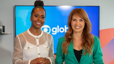 Maju Coutinho e Poliana Abritta sorriem juntas à frente do logotipo da Globo, Maju usa camisa branca translúcida e Poliana veste blazer verde vibrante