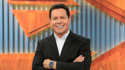 Marcão do Povo sorri com braços cruzados no estúdio do Primeiro Impacto durante apresentação do jornal no SBT