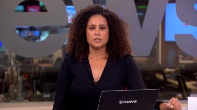Marcelly Setúbal olha para a câmera com expressão séria no estúdio da GloboNews, tem cabelos cacheados volumosos, veste blusa preta e usa notebook