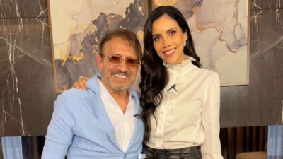 Marcelo de Nóbrega de blazer azul claro e óculos de sol ao lado de Daniela Albuquerque de blusa branca com babados, sorridentes em cenário decorado
