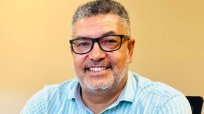 Marcelo Moreira, diretor do canal Veja+, homem de óculos e barba grisalha, veste camisa listrada azul e branca, sorri em frente a um fundo neutro claro