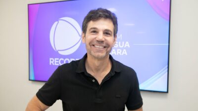 Marcio Atalla sorri de camiseta polo preta em frente a uma tela com o logotipo da Record, com cabelos curtos e grisalhos em um ambiente claro