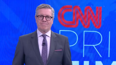 Márcio Gomes veste terno cinza e gravata roxa no cenário do CNN Prime Time com logotipo da emissora ao fundo em tons de azul e vermelho vibrantes
