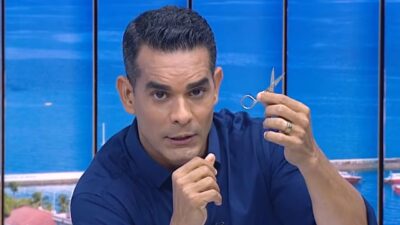 Apresentador Marcus Pimenta em estúdio do Balanço Geral BA na Record Bahia segura pequena tesoura prateada com expressão séria diante de fundo azul