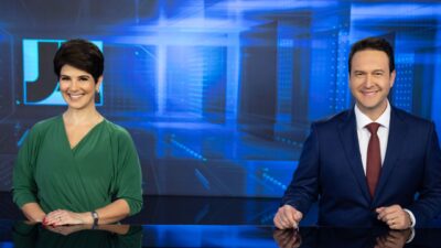 Mariana Godoy de blusa verde e Eduardo Ribeiro de terno azul e gravata bordô sorriem na bancada do Jornal da Record com fundo tecnológico azulado
