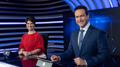 Eduardo Ribeiro veste terno azul com gravata de poá e Mariana Godoy usa blusa vermelha na bancada do Jornal da Record em cenário com luzes neon azuis