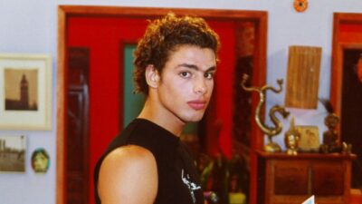 Maumau interpretado por Cauã Reymond em cena de Malhação 2002 usando camiseta preta sem mangas dentro de casa com porta vermelha e móveis ao fundo