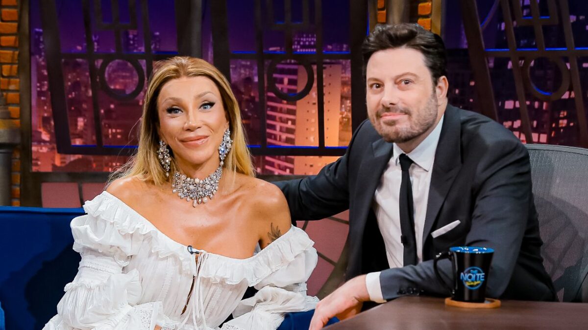 Maya Massafera com vestido branco ombro a ombro e colar prateado ao lado de Danilo Gentili no estúdio do The Noite no SBT