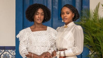Erika Januza como Niara e Duda Santos como Alika, com vestidos brancos de época e rendas, diante de porta azul na novela A Nobreza do Amor