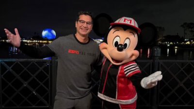 Paulo Antunes sorridente de óculos e camiseta cinza da ESPN ao lado do Mickey Mouse de boné e jaqueta esportiva no Epcot com o Spaceship Earth ao fundo