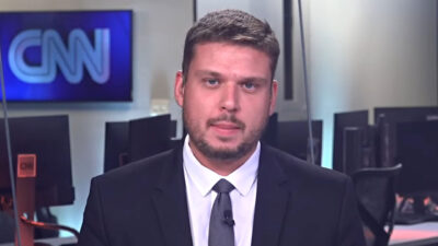 Pedro Teixeira de terno preto e gravata cinza olha para a câmera em estúdio da CNN com bancadas de computadores e monitor exibindo o logotipo ao fundo