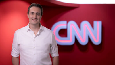 Rafael Furlanetti sorri em estúdio, o executivo veste camisa social branca com mangas dobradas, ao fundo o logotipo da CNN em destaque sobre parede vermelha