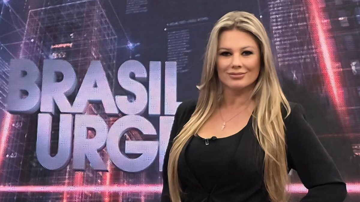 Sandra Redivo posa sorridente à frente do cenário do programa Brasil Urgente, tem longos cabelos loiros, veste blusa e blazer pretos