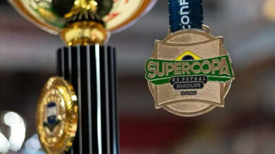 Medalha dourada da Supercopa de Futsal Masculino 2025 pendurada por fita azul ao lado de um troféu com detalhes em dourado e preto em fundo desfocado