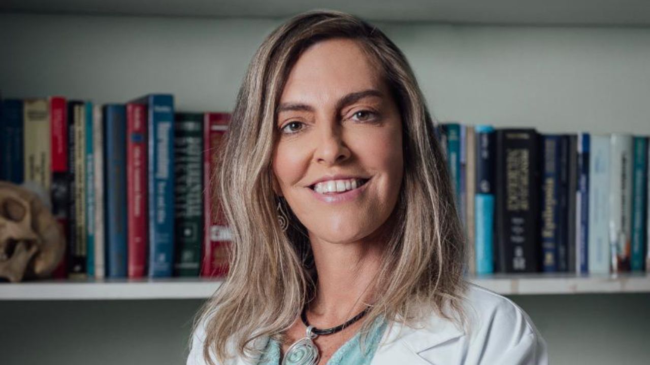 Tatiana Sampaio sorri de jaleco branco e colar, posicionada à frente de uma estante repleta de livros técnicos, representando a área da saúde