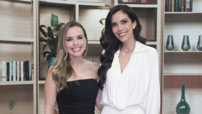 Thaeme Mariôto de vestido preto e Daniela Albuquerque de blusa branca sorriem juntas à frente de uma estante com livros e vasos em estúdio de TV