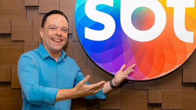 Thiago Gardinali sorri apontando para o logotipo colorido do SBT, veste camisa azul clara e está posicionado em frente a uma parede de madeira clara