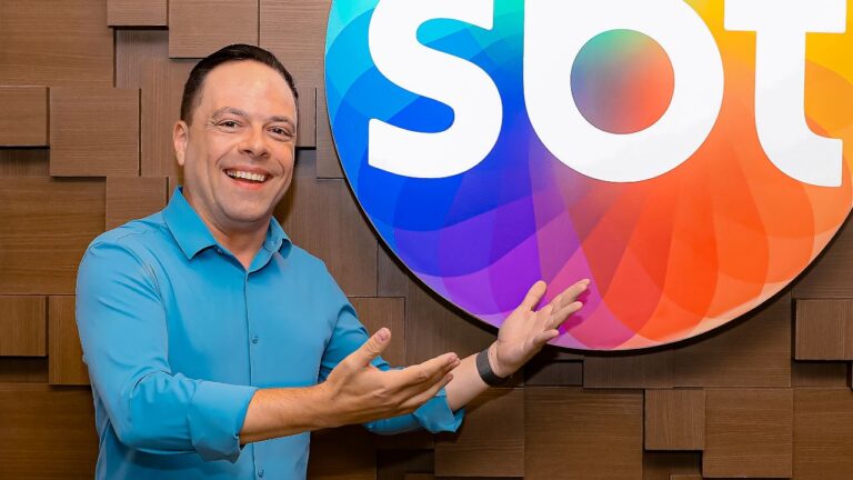 Thiago Gardinali sorri apontando para o logotipo colorido do SBT, veste camisa azul clara e está posicionado em frente a uma parede de madeira clara