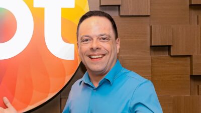 Thiago Gardinali sorri de frente vestindo camisa azul clara, ao fundo parede de madeira com relevos quadrados e parte do logotipo colorido do SBT