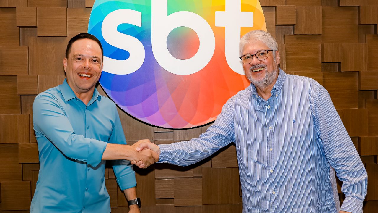 Thiago Gardinali e Leon Abravanel sorriem e apertam as mãos em frente ao logotipo colorido do SBT, ambos vestem camisas sociais em tons de azul