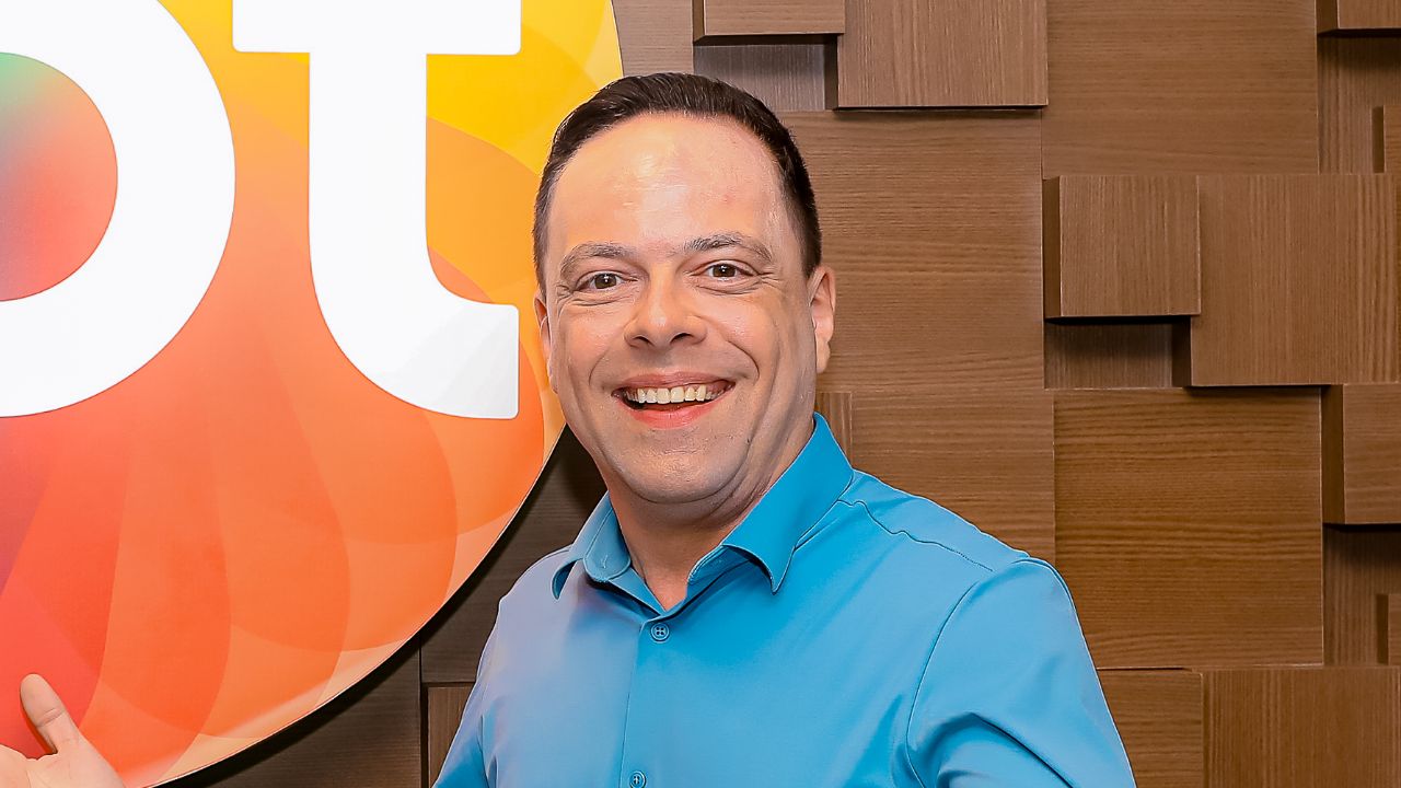 Thiago Gardinali sorri de frente vestindo camisa azul clara, ao fundo parede de madeira com relevos quadrados e parte do logotipo colorido do SBT