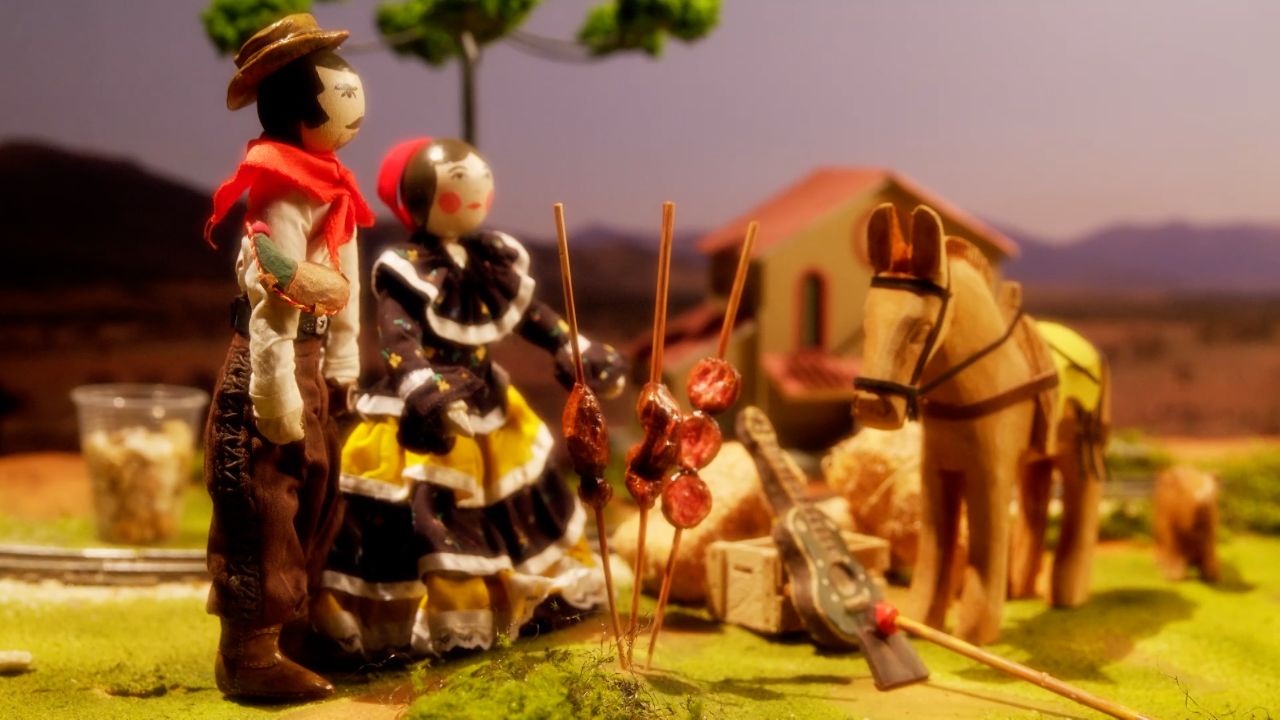 Maquete com casal caipira ao lado de cavalo e violão em cenário rural com espetinhos sobre fogueira e casa ao fundo
