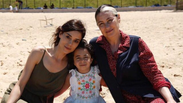 Zeynep, Melek e Gonul, personagens da novela Mãe, sentadas na areia de uma praia sob o sol, a menina Melek está ao centro entre as duas mulheres