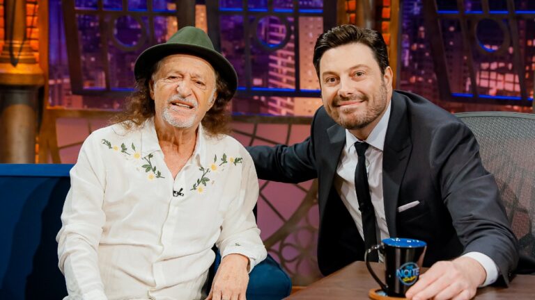 Alceu Valença veste camisa branca bordada com flores e chapéu verde ao lado de Danilo Gentili, de terno preto, no cenário do programa The Noite no SBT