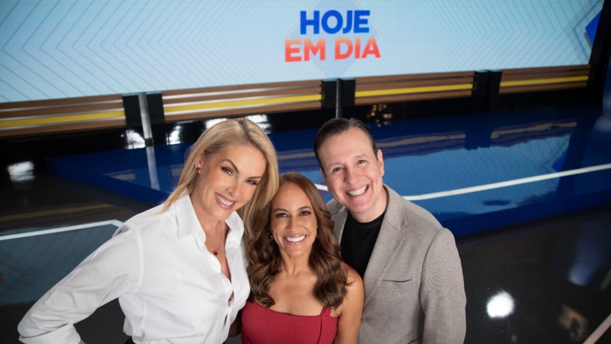 Ana Hickmann, Renata Alves e Celso Zucatelli sorriem abraçados no cenário do programa Hoje em Dia, ao fundo há um telão com o logotipo da atração