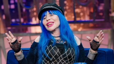 Baby do Brasil sorri com cabelos azuis longos, boina preta, blusa de rede com brilhos e luvas sem dedos, sentada no sofá azul do programa The Noite