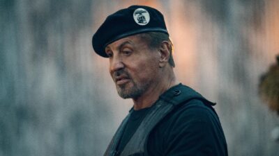 Ator Sylvester Stallone como o personagem Barney no filme Os Mercenários 4, usa boina preta com emblema de caveira e colete tático em cena de ação