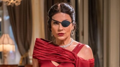 Flávia Alessandra, como a vilã Sandra na novela Êta Mundo Melhor!, usa tapa-olho preto, vestido de gala vermelho e colar de brilhantes em cenário luxuoso