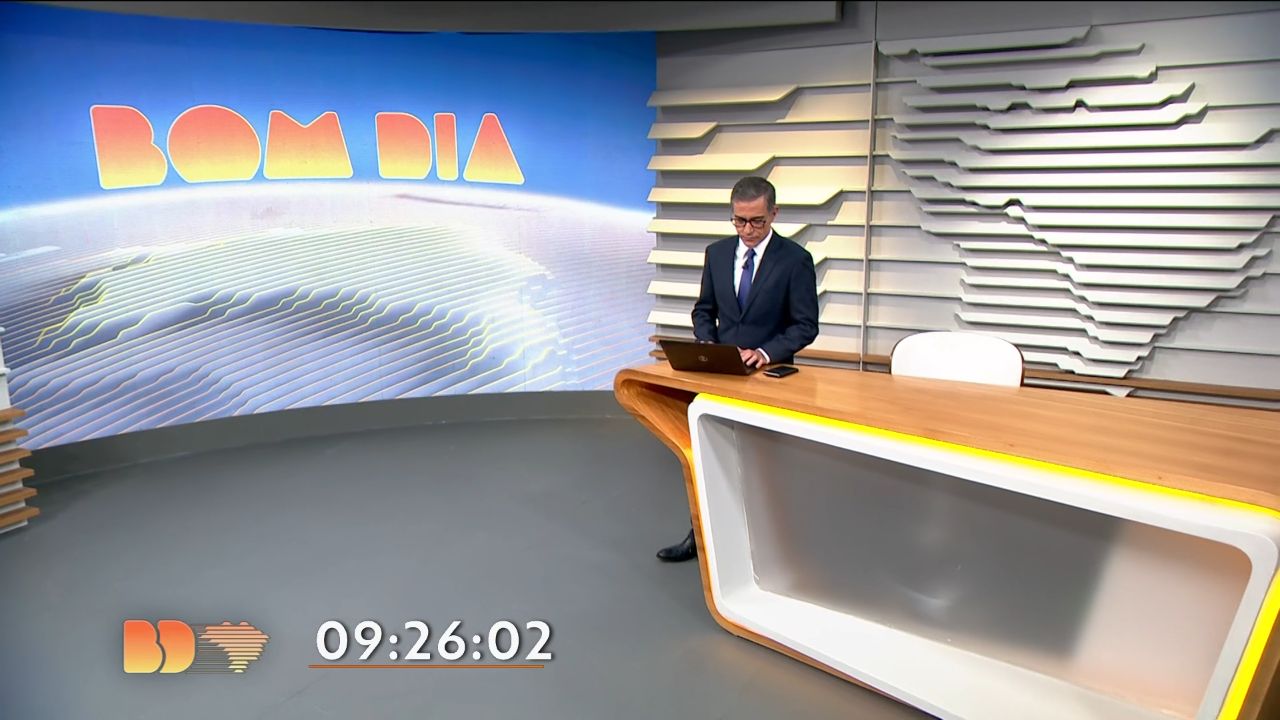 Hélter Duarte em pé no cenário do Bom Dia Brasil, usa notebook sobre bancada de madeira, ao fundo telão azul com logotipo do telejornal da Globo