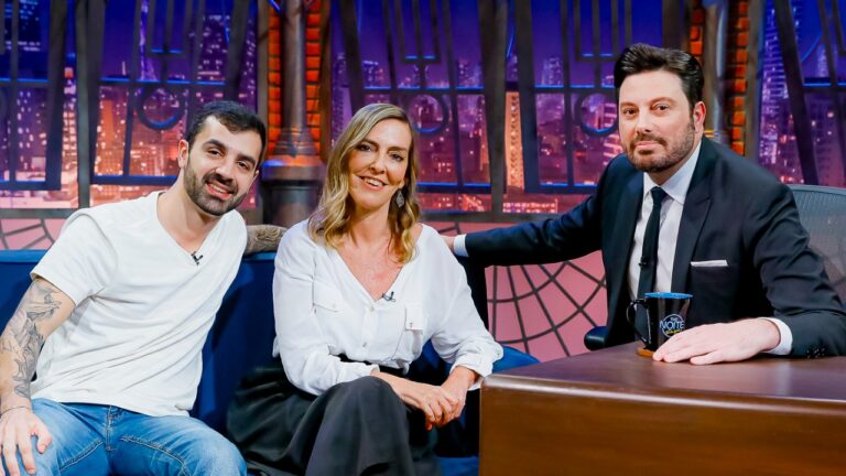 Bruno Drummond de camiseta branca e Tatiana Coelho sorriem ao lado de Danilo Gentili no sofá azul do The Noite, no SBT, com a cidade iluminada ao fundo
