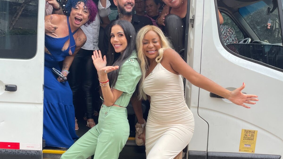 Cariúcha e Daniela Albuquerque posam sorrindo na porta de van ao lado de grupo de pessoas durante gravação externa