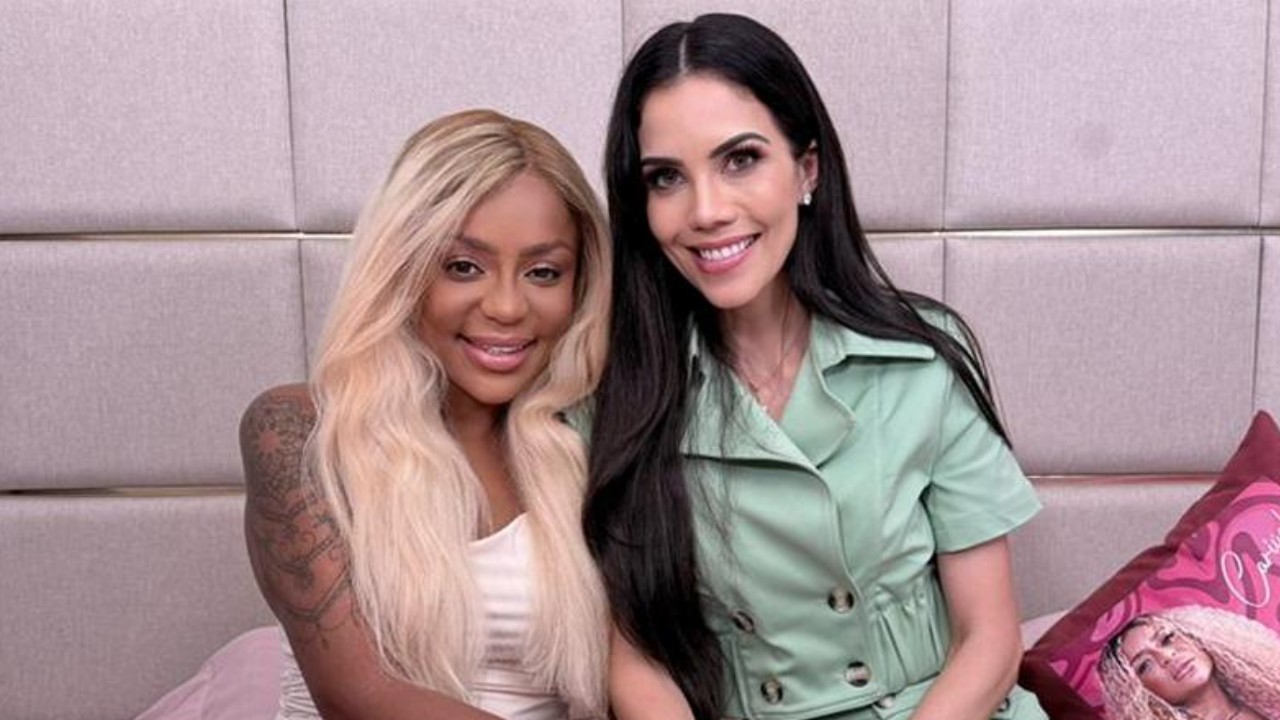 Cariúcha e Daniela Albuquerque sentadas lado a lado sorrindo em ambiente interno com parede acolchoada ao fundo