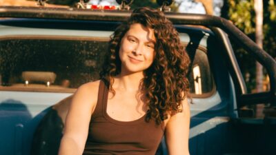 Carolina Belarmino como Mariana sorri em frente a caminhonete ao ar livre em cena do filme DOMA com luz de fim de tarde