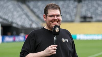 Casimiro Miguel de camiseta preta e óculos, sorri enquanto segura microfone da CazéTV com espuma personalizada, em um estádio de futebol vazio
