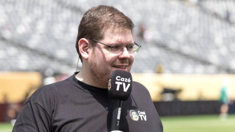 Casimiro Miguel de camiseta preta e óculos, segura microfone da CazéTV com espuma personalizada, em um estádio de futebol com arquibancadas ao fundo
